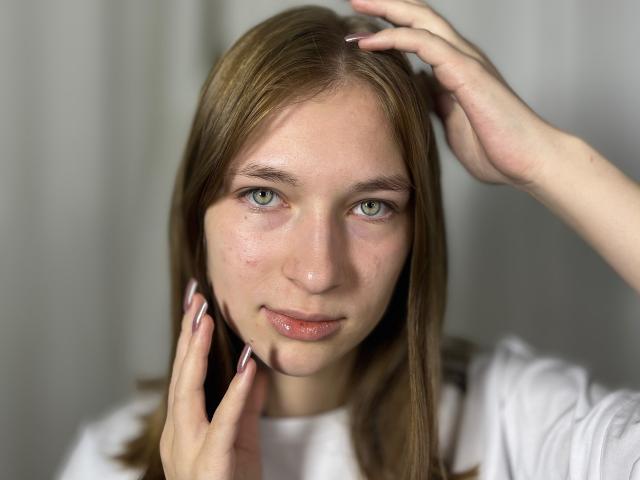 Fotografia ravvicinata di BeaDealba, una giovane donna con la pelle chiara, gli occhi verdi e i capelli castani, che si tocca la testa con una mano. e il suo viso con l'altra, indossando una camicia bianca. Tensione sessuale.