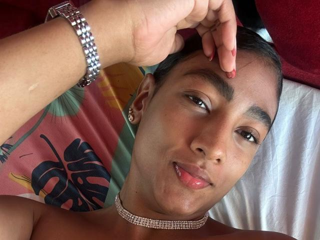 Close-up of NatalyBlonder with tan skin, dark hair, and a slight smile, wearing a silver choker and bracelet, touching her head with a finger, lips slightly parted, looking seductive.