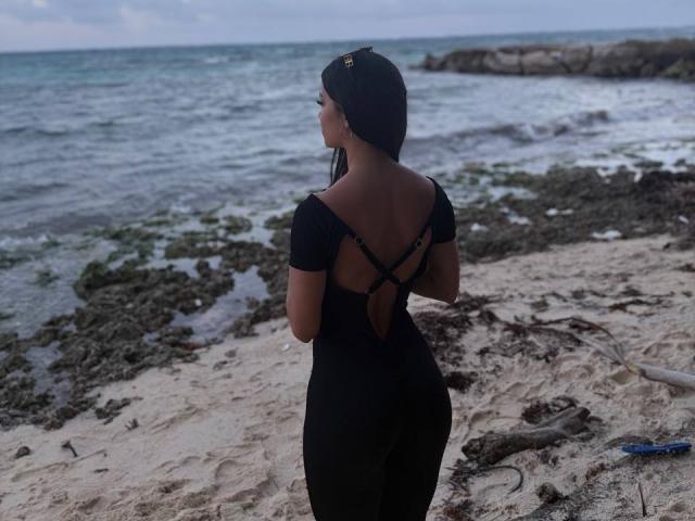 LaurentRay está de pie en una playa rocosa, mirando hacia el océano. Lleva un vestido negro con la espalda descubierta, y sus trenzas le caen sobre los hombros. Ella está mirando hacia otro lado, con el pene ligeramente visible..