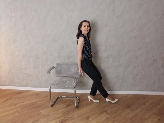 DaisyEdit, with curly brown hair, leans against a gray textured wall, wearing a black polka dot top, black pants, and white heels, standing beside a striped chair on wooden floors.