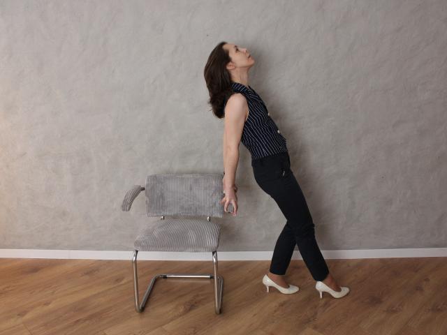 DaisyEdit leans against a gray, wire-frame chair with her back arched, head tilted back, wearing a black polka-dot top and black pants, with white heels, on a wooden floor.