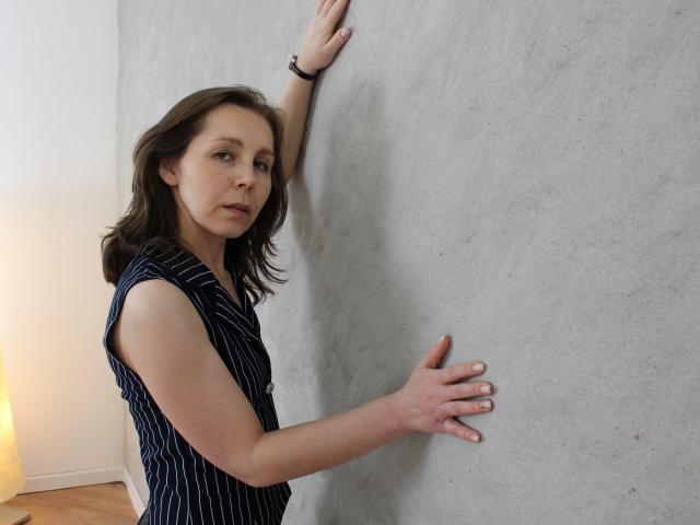 Daisy, con capelli castano di media lunghezza, indossa una maglietta a righe nere e bianche senza maniche e si appoggia a una superficie grigia con una texture particolare. un muro con una mano alzata e l'altra tesa, che guarda leggermente di lato.