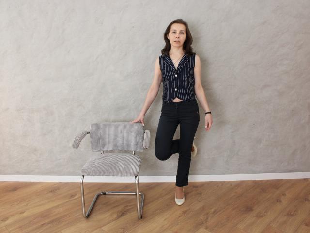 DaisyEdit leans against a gray textured wall, wearing a black and white striped sleeveless top and black pants, touching a gray mesh chair with her right hand. Wooden floor. Fucking casual pose.