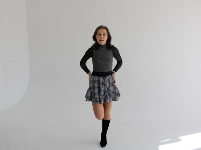 DaisyEdit staat zelfverzekerd in een witte studio, gekleed in een zwart topje met lange mouwen van kant en een zwart-wit gestreept rok, met zwarte enkellaarsjes. Ze kijkt recht in de camera, met haar handen op haar heupen..