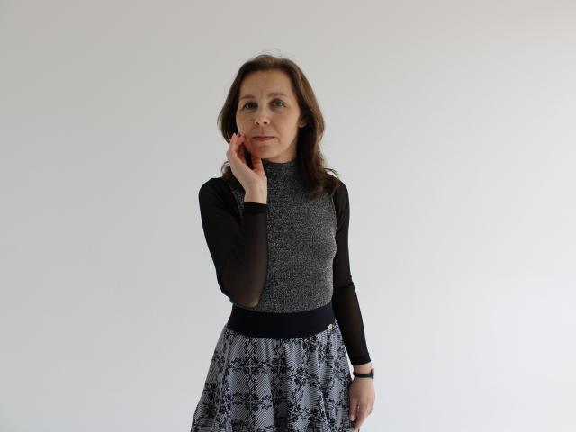 DaisyEdit, una mujer de mediana edad con cabello castaño ondulado, está de pie frente a un fondo blanco. Ella lleva una blusa negra de manga larga con un corpiño gris brillante y una falda estampada. Ella toca su mejilla con la mano derecha, mirando ligeramente hacia un lado. Su expresión es neutra..