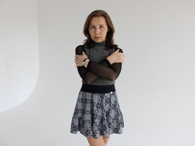 DaisyEdit está em frente a uma parede branca, com os braços cruzados, vestindo uma blusa preta transparente e uma saia com padrão azul e branco. Ela tem cabelo castanho escuro e uma expressão séria. Uma expressão muito séria..