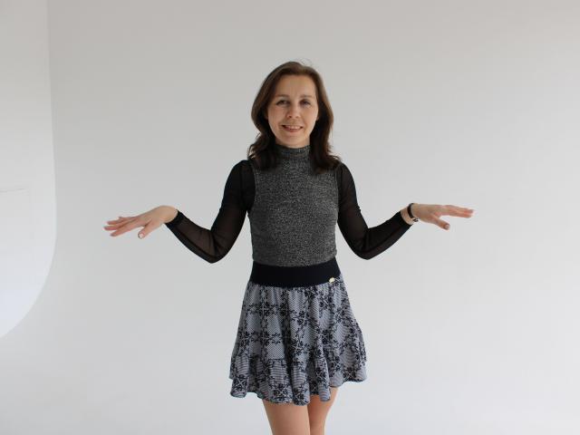 DaisyEdit stands center-frame, arms outstretched, wearing a black long-sleeve top and black-and-white patterned skirt. She has curly brown hair, a watch on her left wrist, and a subtle smile.