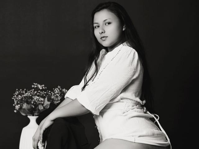 Black-and-white photo of CorinaHahn, sitting with legs apart, wearing a white shirt and skirt, long dark hair, looking over shoulder, hand resting on vase with small flowers. Fucking sexy pose.