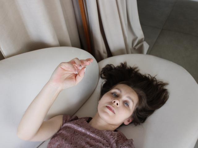 DaisyEdit, with disheveled dark brown hair, lies on a white chair, wearing a shimmering brown top, hand raised, blue eyes looking up, soft light, grey tile floor, beige curtains.