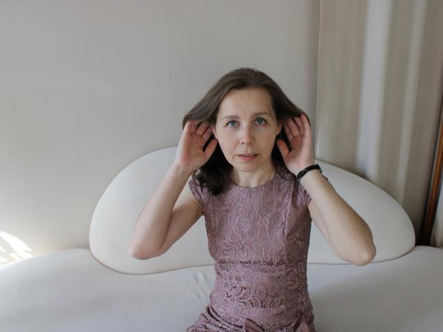 Daisy, mit braunen Haaren, trägt ein eng anliegendes, glitzerndes rosa Kleid und sitzt auf einem weißen Sofa, wobei sie ihre Hände vor ihren Ohren hält. Sie hat blaue Augen. Blick leicht zur Seite gerichtet, subtile sexuelle Spannung..