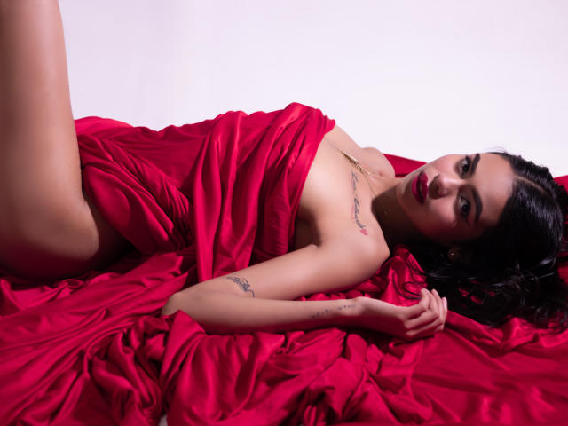 CrystalDavids lies on red, rumpled fabric, nude except for the fabric partially covering her. She gazes at the camera with dark eyes, black hair, tattoos on her arm. Fucking sexy.