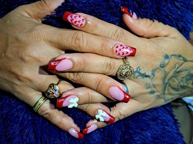 As mãos de Kate Monroe repousam sobre uma superfície escura, semelhante a pele de animal, exibindo unhas longas com pontas vermelhas e rosa, e bolinhas vermelhas. e detalhes em branco. Usando anéis de ouro, tatuagem na mão direita. Incrivelmente sexy..