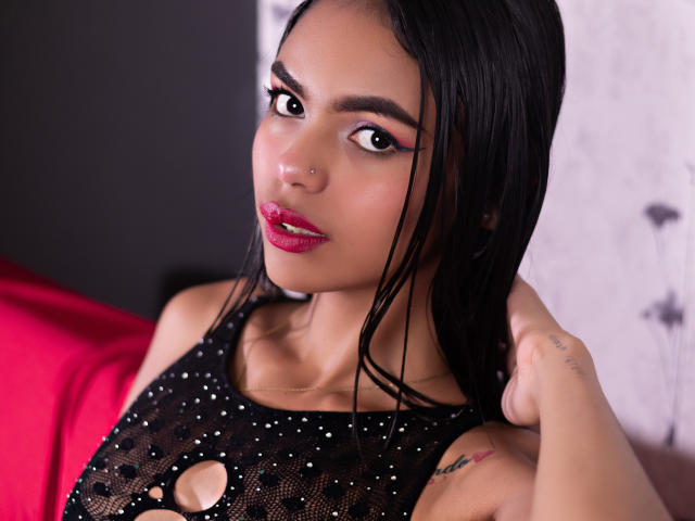 Fotografia ravvicinata di CrystalDavids, una sexy donna latina con capelli neri bagnati, rossetto rosso e un piercing al naso. Indossa una maglia nera in rete con un taglio, e guarda direttamente la fotocamera con uno sguardo seducente..