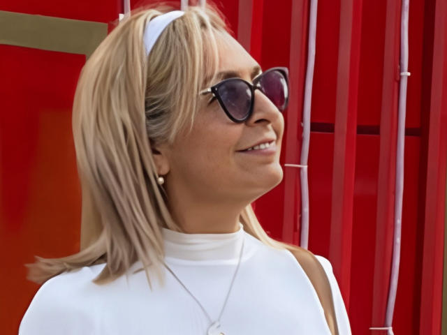 Fotografía de primer plano de CristallMature con cabello rubio hasta los hombros, una diadema blanca, gafas de sol negras y un atuendo blanco. Cuello alto. Barras de metal rojas en el fondo. Cristall, mirando hacia arriba, sonriendo, con una ligera tensión sexual..