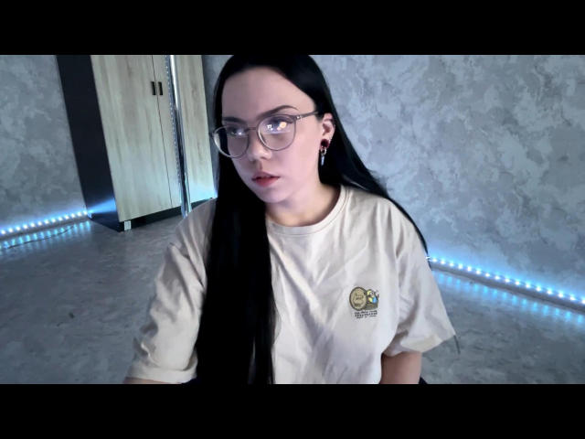 Ghislaine Koplin, donna asiatica con lunghi capelli neri, che indossa occhiali e una maglietta bianca con un piccolo logo, che guarda. Direttamente verso la telecamera, luci al neon blu posizionate a terra, parete in cemento..