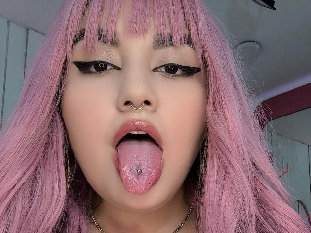 Gros plan de MarlaBriar avec des cheveux roses, un anneau de nez et un eye-liner noir. Elle sort sa langue, qui est percée. Son expression est provocante. L'image est très suggestive sur le plan sexuel..