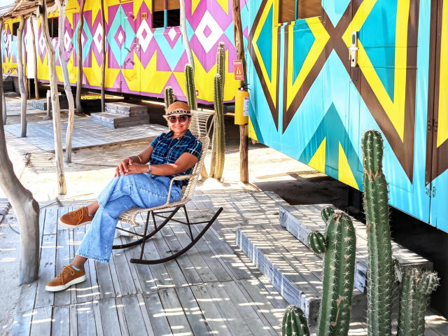 CristallMature, vêtue d'une chemise à pois et d'un jean bleu, est assise dans un fauteuil à bascule noir, sous un ciel éclatant. Des murs avec des motifs géométriques et des cactus. La lumière du soleil projette de nettes ombres sur le plancher en bois..