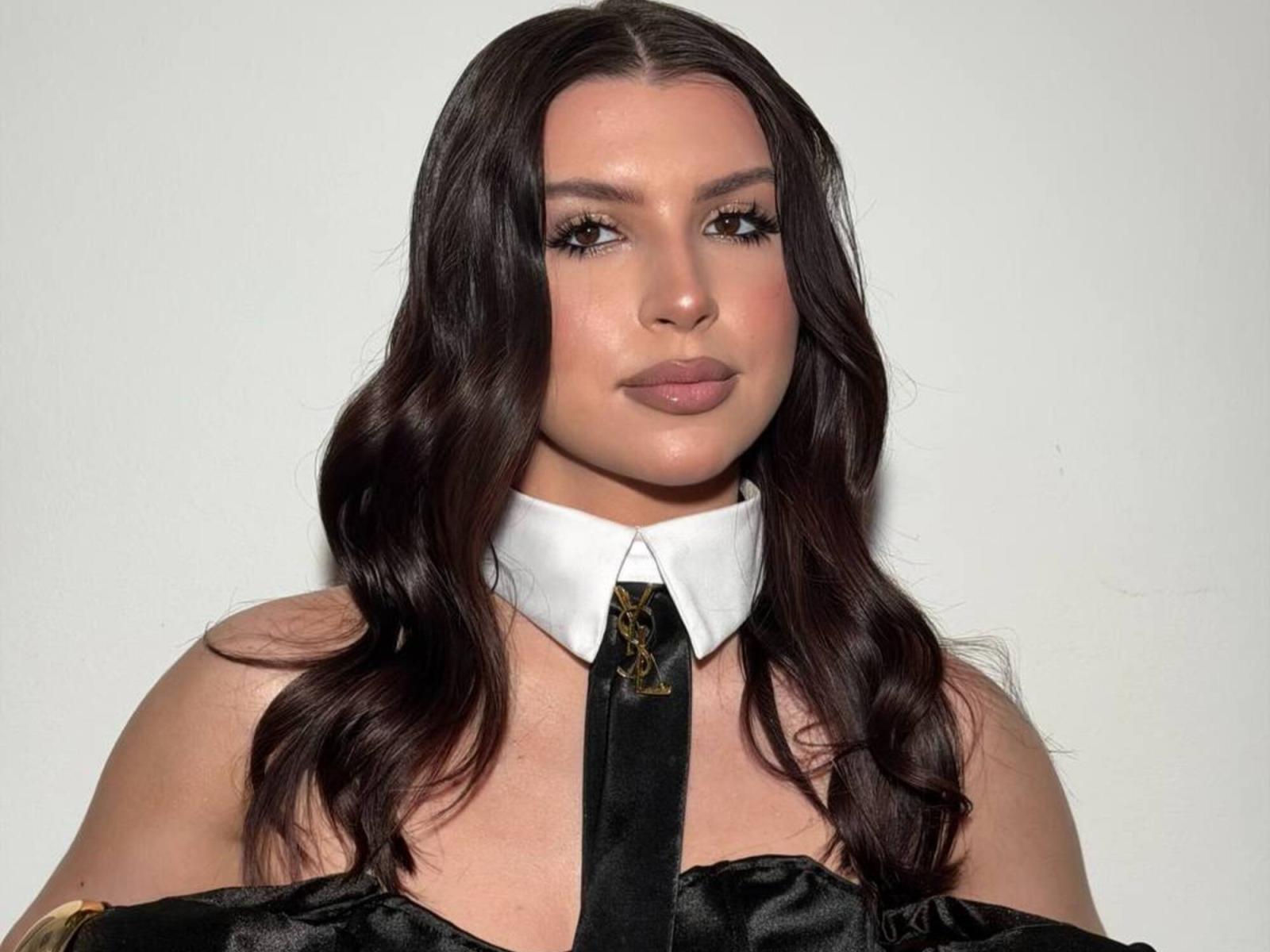 Fotografía de KeenaJaminet, una mujer de curvas con cabello negro largo y ondulado, vestida con una camisa blanca con cuello y una corbata negra. mirando directamente a la cámara con una expresión sutil y sensual.