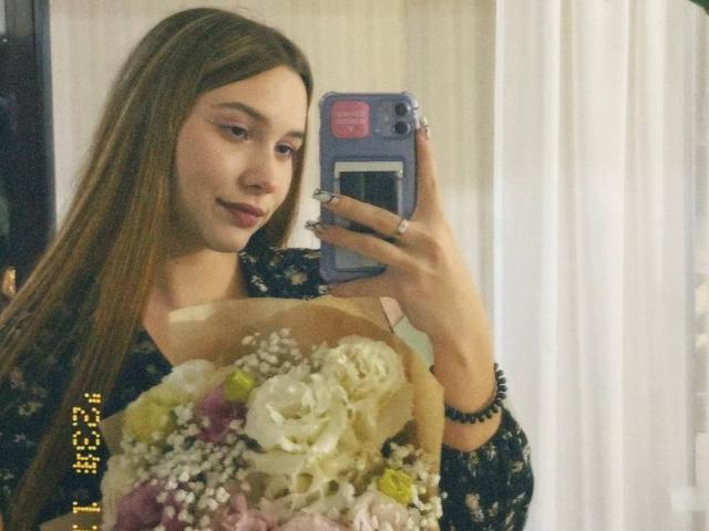 Photographie de Kenisha Haislett, une jeune femme aux longs cheveux bruns, tenant un bouquet de fleurs, prenant un selfie devant un miroir. avec un smartphone. Elle a un sourire discret, et elle porte un haut sombre avec un motif..