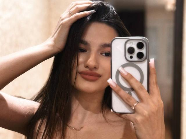 JinnyWalthers, nue, tenant un iPhone blanc avec un support en anneau, une main dans les cheveux, sourire doux, lumière naturelle, cheveux noirs, teint moyen, maquillage discret, décontracté, sexy, confiant, autoportrait en gros plan..