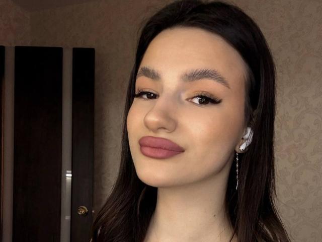 Gros plan de JinnyWalthers, une jeune femme aux longs cheveux noirs, à la peau claire et aux lèvres rouges pulpeuses. Elle regarde légèrement sur le côté, avec une expression confiante, et porte des boucles d'oreilles argentées. Tension sexuelle.