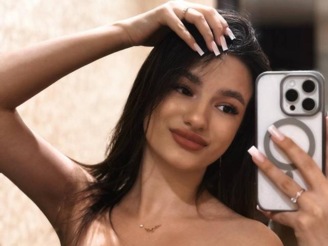 Fotografia di JinnyWalthers che tiene in mano uno smartphone con una custodia argentata con la mano destra e si sta scattando un selfie. Ha i capelli scuri, la pelle color nocciola, le unghie lunghe e bianche e un sorriso delicato. La sua mano sinistra è appoggiata sulla testa. È senza top, e si può vedere una piccola collana d'oro. Molto attraente..