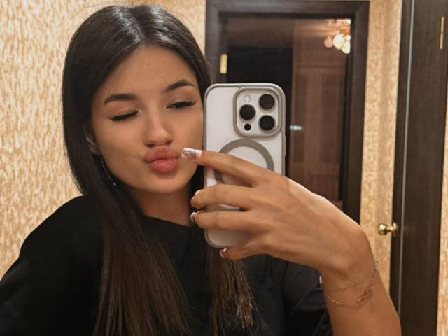 JinnyWalthers with long black hair, pouting lips, and a sexy expression, holds a silver iPhone with one hand, wearing a black shirt and gold bracelet, in a bathroom. Fucking hot.