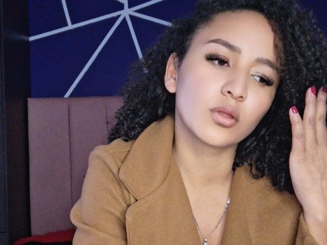 Photograph of AbvrilDubois with light brown skin and curly black hair, wearing a tan coat, red nail polish, and a silver necklace. She's looking away with a slightly pouty expression.