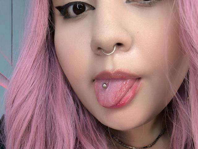 Primer plano del rostro de MarlaBriar, enfocándose en su cabello rosa, su aro de plata en la nariz y su lengua con un piercing de plata. Sus ojos están medio cerrados, los labios entreabiertos de forma seductora, mostrando la lengua rosada..