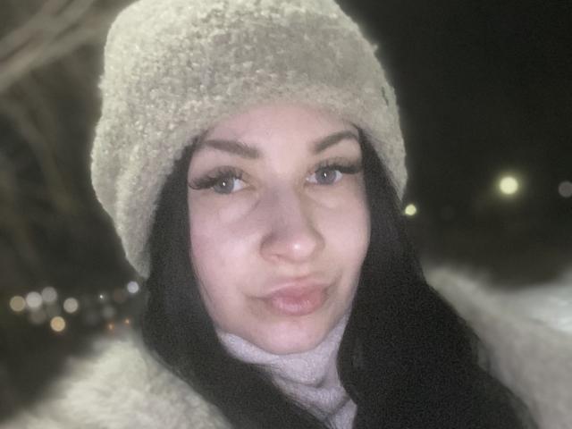 Primo piano di StarrySiren, una giovane donna con la pelle chiara e i capelli neri, che indossa un cappello e una sciarpa bianchi di lana, e che sembra irradiante. Una luce notturna con un sorriso dolce e invitante. Luci bokeh sullo sfondo.