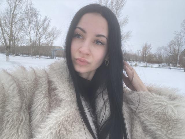 StarrySiren, met lang zwart haar, draagt een pluizige witte bontjas en poseert buiten in een besneeuwd landschap. Ze kijkt recht in de camera, haar hand in haar haar, met een koude, winterse sfeer..