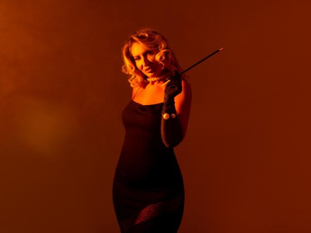 GinaBear with curly blonde hair stands in a red-orange light, holding a conductor's baton. She's wearing a tight black dress with fishnet pattern on her right thigh. Sexual tension is high.