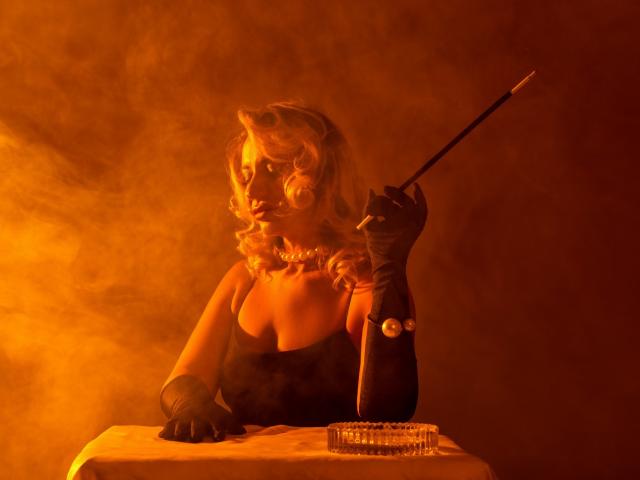 GinaBear with curly blonde hair, wearing a black strapless top and long gloves, holds a cigarette holder in her right hand. Smoky orange light highlights her sultry, confident pose. Cigarette pack on table.