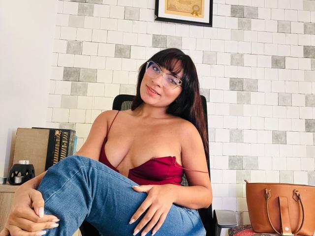 Ameline Rousseau, com cabelo castanho escuro e óculos, sentada em uma cadeira, vestindo uma blusa vermelha sem alças e jeans azuis, pernas bolsa marrom, com alça, posicionada à direita, parede de azulejos ao fundo..