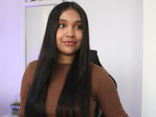 Fotografía de BelleLane con cabello largo y negro, una blusa marrón de manga larga y una sonrisa sutil. Sus ojos marrones miran ligeramente hacia arriba, irradiando una sutil tensión sexual. Fondo blanco brillante.