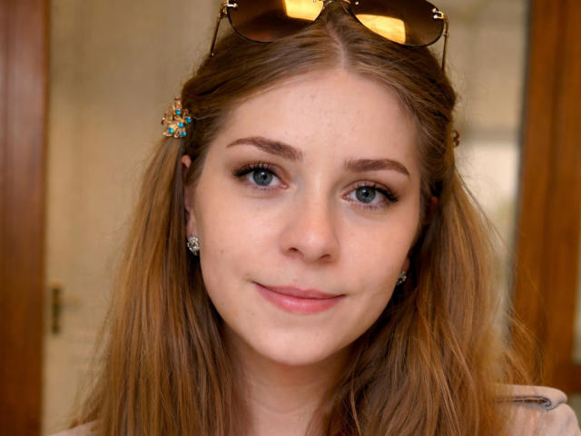 Foto em close-up de SoftlySabrina, uma jovem com pele clara, olhos azuis e cabelos castanhos longos, usando óculos de sol. Um lenço na cabeça, um prendedor de cabelo colorido e brincos de diamante. Sorrindo levemente.