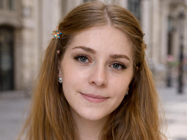 Nahaufnahme von SoftlySabrina mit heller Haut, blauen Augen und hellbraunen Haaren, die eine farbenfrohe Haarnadel und einen Diamanten trägt. Ohrringe. Unschärfer Hintergrund einer Stadt, sanftes Lächeln, leichte sexuelle Spannung.