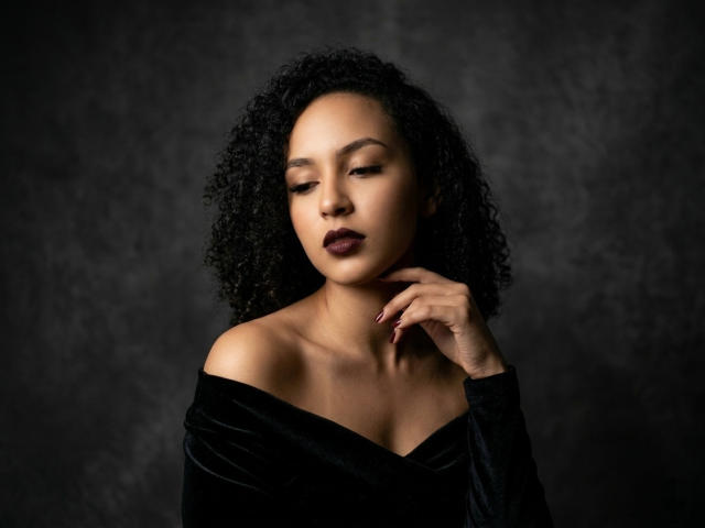 Fotografía de AbvrilDubois con cabello negro rizado, vistiendo una blusa negra con un hombro descubierto, ojos cerrados, labios ligeramente entreabiertos. mano cerca del cuello, maquillaje oscuro, iluminación oscura, mirada intensa, ambiente sensual..