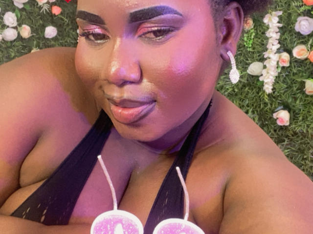 Fotografia ravvicinata di Dilay, una donna nera formosa con un trucco viola e rosa brillante, che guarda in basso con un leggero sorriso. Indossa un top nero con spalline che lascia intravedere molto il décolleté. Lo sfondo presenta luci bianche e rosa sfocate..