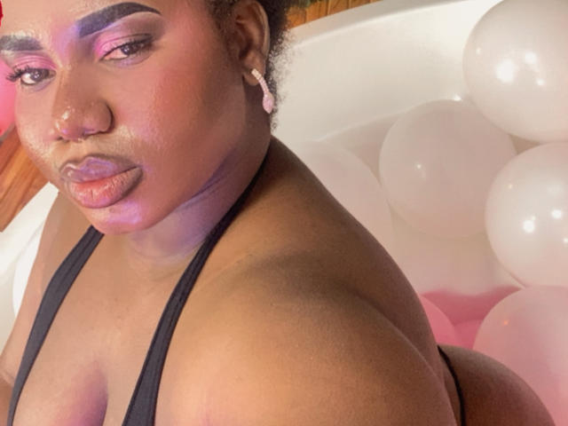 Primer plano de Dilay, una mujer negra de curvas, con una piel radiante, sombra de ojos morada y lápiz labial rosa, vestida con un top de tirantes negro. arriba, rodeada de globos blancos, mirando directamente a la cámara.