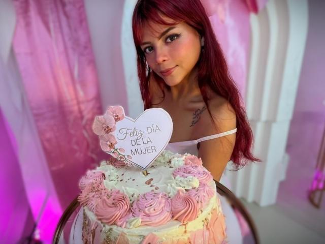 ViioletteRoselle, con cabello rojo largo, sosteniendo un letrero blanco con forma de corazón, adornado con flores rosas y la inscripción "Feliz cumpleaños Rosmery". escrito en él. Ella lleva un top sin tirantes y sostiene un ramo de flores rosa y blanco..