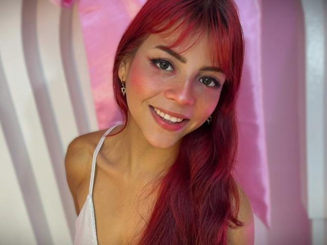 Close-up de ViioletteRoselle, uma mulher atraente com longos cabelos ruivos, sorrindo, vestindo uma camiseta branca, rosa e branca. contexto. Seus olhos azuis e o leve rubor adicionam charme. Incrivelmente fofa..