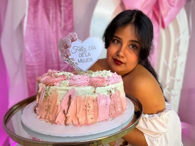 Cassandra Valmont mit langen schwarzen Haaren, rotem Lippenstift und einem weißen Top mit Schulterpartie, die einen rosa Kuchen mit Zuckerguss hält. "Carita de Dios" auf einem weißen Schild. Sexuelle Spannung durch ihren direkten Blick..