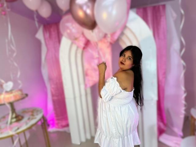 Cassandra Valmont staat in een kamer met roze en witte ballonnen, gekleed in een witte jurk met een open schouder, en kijkt om. Verleidelijke uitdrukking. Witte boog en roze gordijnen op de achtergrond. Verdomd sexy..