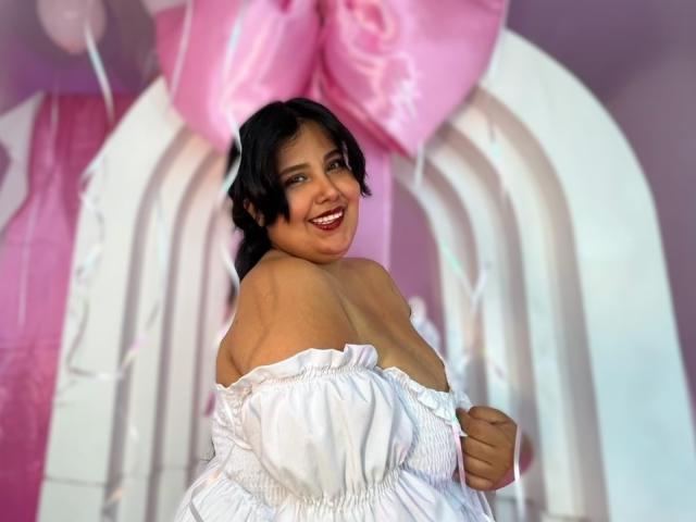 Cassandra Valmont, con i capelli ricci e scuri, sorridente, indossa un abito bianco con una spalla scoperta, tiene in mano del tessuto e ci sono palloncini rosa. sfondo, tensione sessuale, attraente, scollatura visibile, illuminazione intensa, posa giocosa.