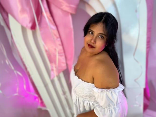 Cassandra Valmont, con cabello negro largo, lápiz labial rojo oscuro y una blusa blanca con un hombro descubierto, mirando hacia atrás con una expresión seductora. sonríe. Tela rosa y blanca drapeada en el fondo. Increíblemente sexy..