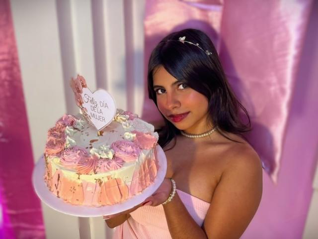 CelesteMorel holds a pink-iced birthday cake with "Happy Birthday" on a white card, wearing a strapless pink dress, pearl necklace, and red lipstick. Bright pink and white background. Sexual tension.