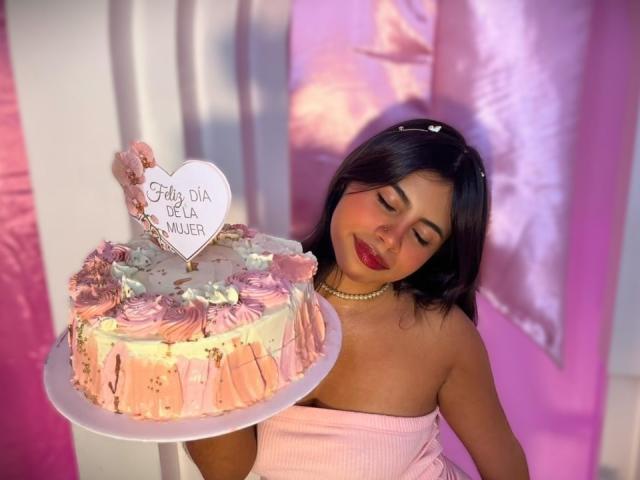 Fotografía de Celeste Morel sosteniendo un pastel cubierto de glaseado rosa con decoraciones de estrellas blancas y un letrero con forma de corazón. Leyendo "Happy Day Real Girl". Ella está sonriendo, vestida con un vestido rosa sin tirantes..