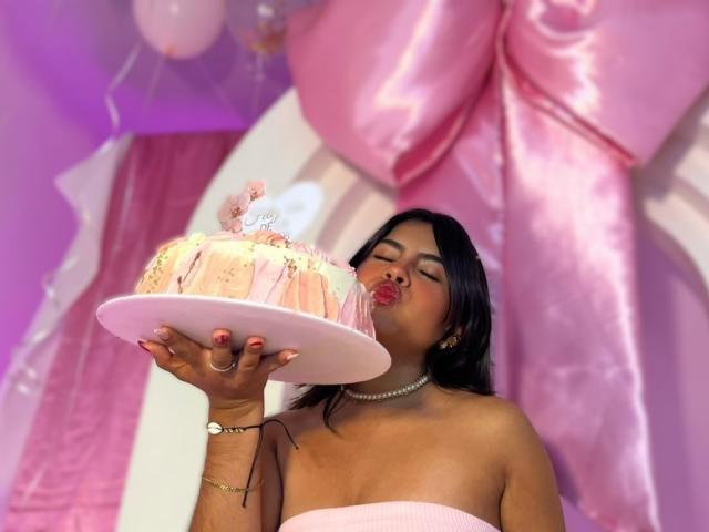 CelesteMorel, topless, holding a frosted cake with pink frosting, licking it, dark hair, pearl necklace, pink balloon backdrop, bright lighting, sexy, cake in hand, erotic tension, close-up, vibrant colors.