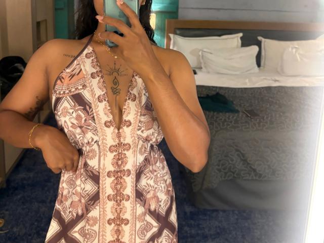 Secretlysweet-hot draagt een bruin-witte jurk met een haltertop, met een hand voor het gezicht, een tatoeage op de borst, en een gouden armband. Staand voor een bed met witte kussens. Seksuele spanning. Slaapkamer setting..